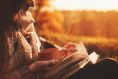 Eine junge Frau liest in Abend- oder Morgendämmerung mit einer dampfenden Tasse in der Hand ein Buch und blättert gerade die Seite um.