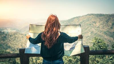 Eine Frau mit roten Haaren ist von hinten zu sehen und sie breitet vor einer bergigen Landschaftskulisse eine Landkarte aus, die sie vor sich hält. Sie steht vermutlich auf einer Aussichtsplattform.